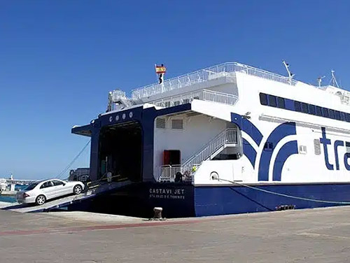 Un ferry de la compañía Trasmapi en el puerto, con un coche blanco entrando a bordo por una rampa. El ferry, llamado Castavi Jet, está preparado para transportar vehículos y pasajeros. El cielo es azul y despejado.