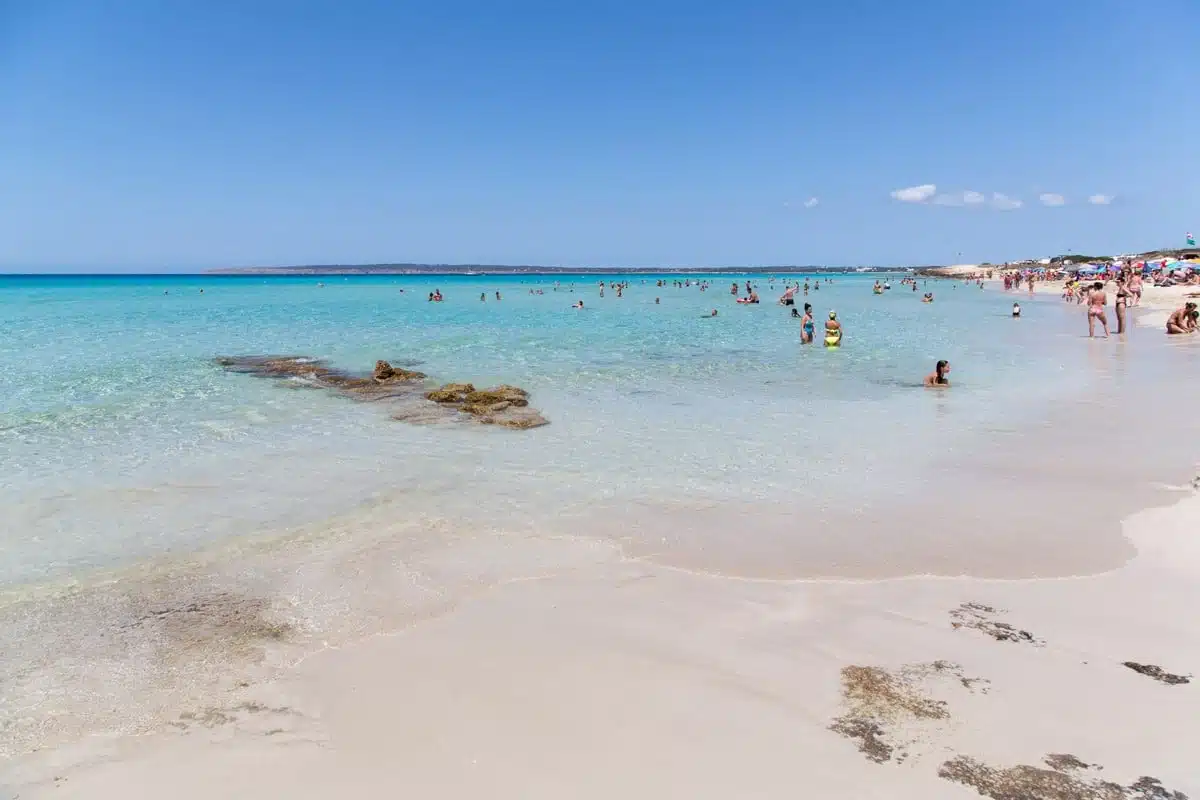 Banyistes a l’aigua turquesa i poc profunda de la platja d’es Arenals, a Formentera, amb sorra blanca, unes roques a la vora i para-sols al fons