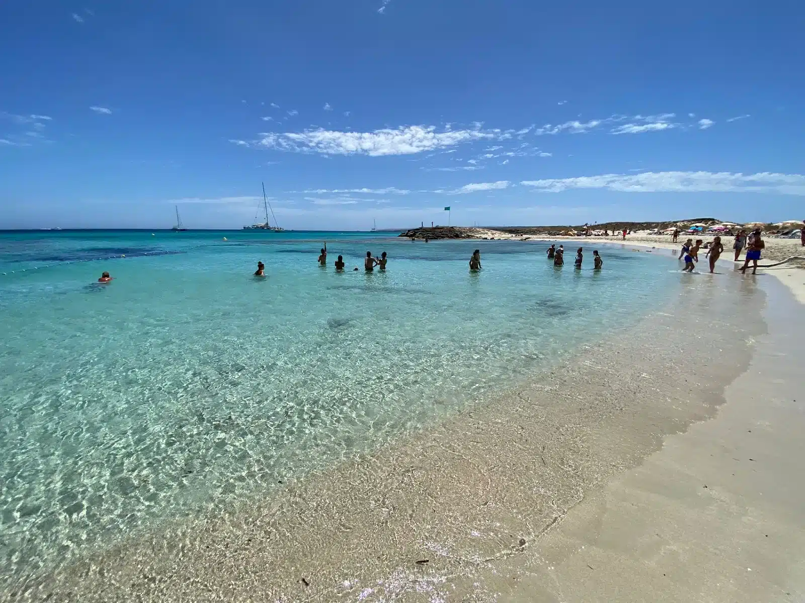 Badegäste im türkisfarbenen Wasser nahe dem Ufer am Strand Llevant auf Formentera, im Hintergrund eine grüne Flagge und Segelboote